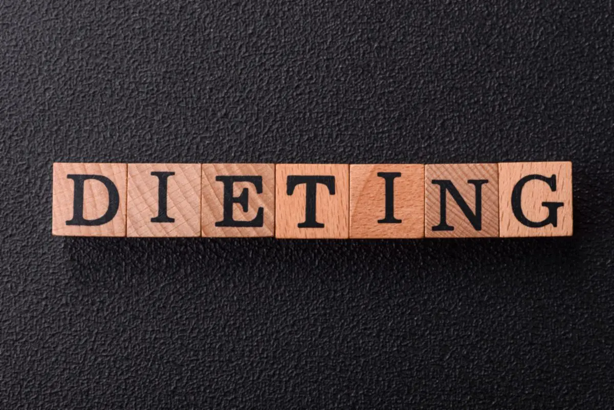 Wooden letter blocks spelling 'DIETING' laid out on a dark textured surface (image emphasizes dieting).