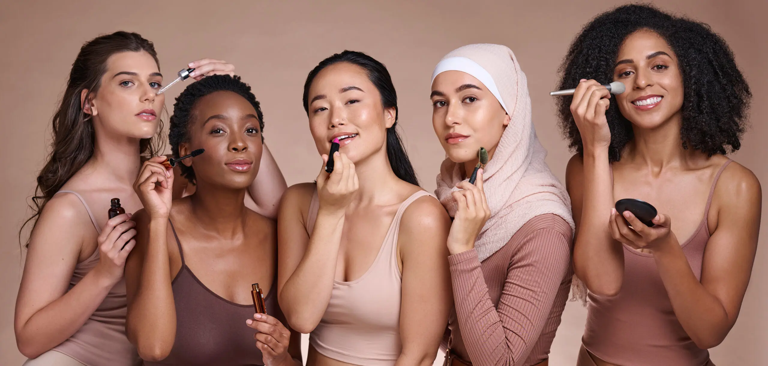 Five diverse women applying makeup together, holding brushes and lipstick against a neutral tan background.
